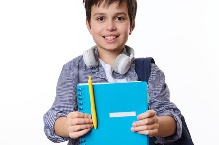 Adorable teen boy, primary school student holding out a blue copybook and yellow pen, smiling a cute...