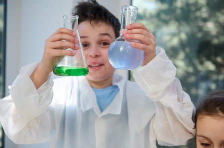 Details: selective focus on the hands of a schoolboy in white lab coat, holding lab flasks with...