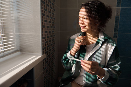 Positive middle aged woman holding inkjet pregnancy test in home bathroom, looking nervously out the...