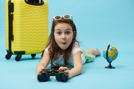 Happy and surprised little traveler child girl with binoculars, expressing positive emotions and surprise, looking at camera, lying near a yellow suitcase and globe, isolated on blue color backgroundの写真素材