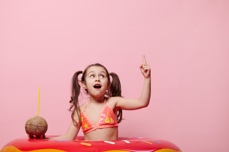Caucasian cute little child girl, wearing stylish bright swimsuit, pointing index finger at copy space for ads, isolated on pink background, looking up while posing with coconut drink and swim ringの写真素材