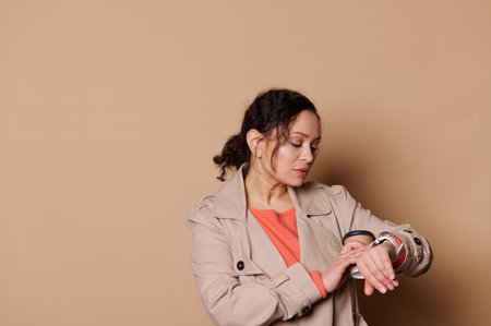 Confident multi-ethnic woman in beige coat, entrepreneur checking time on her wrist watch, harrying...