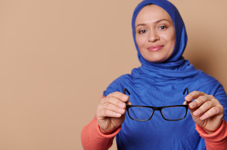 Selective focus on black spectacles - eyeglasses in the hands of a blurred stylish Arab Muslim in blue hijab, smiling looking at camera, isolated on beige background. Health Fashion and ophthalmologyの写真素材