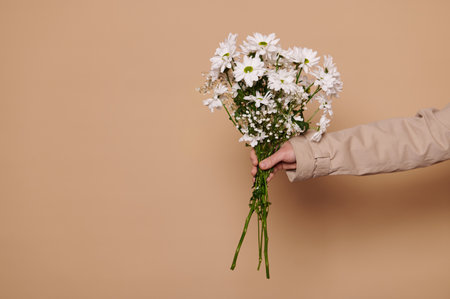 Close-up of hand, holding out over beige background a bunch of chrysanthemum and chamomile flowers. Beautiful spring bouquet of fresh flowers for Womens Day, birthday or other festive event, occasionの写真素材