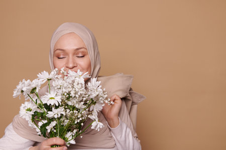 Close-up face portrait of a beautiful Muslim woman in a beige hijab, sniffing a bouquet of white chamomile flowers on isolated beige background with copy ad space. International Womens Day conceptの写真素材