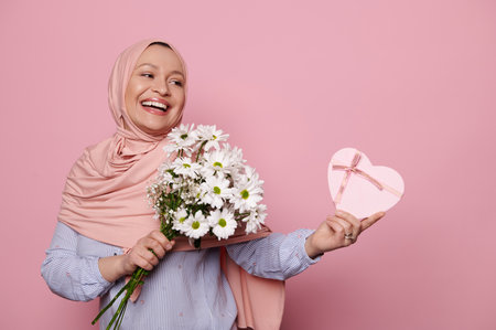 Cheerful Arab Muslim woman in pink hijab, with a beautiful flower bouquet and a happy present in a heart shaped gift box, smiling looking aside at copy ad space, isolated pink background. Womens Dayの写真素材