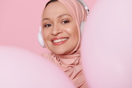 Close-up portrait of middle aged Muslim woman wearing headphones and pink hijab, smiling with beautiful toothy smile, peeking out from behind pink pastel inflatable balloons, isolated pink backgroundの写真素材