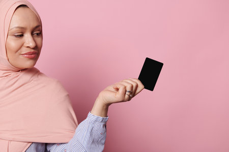 Confident attractive Arab Muslim woman with head covered in pink hijab, holding a black plastic bank credit card over isolated pink background. People. Finance. Business. Mobile banking. Copy ad spaceの写真素材