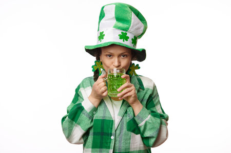 Pretty woman dressed in carnival party hat with clover leaves and green checkered shirt, looks at camera, makes sip of green beer, celebrating Saint Patricks Day, March 17, isolated white backgroundの写真素材