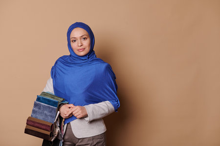 Professional portrait of a Middle-Eastern Muslim woman in formal clothes and blue hijab, interior designer holding upholstery fabric samples, looking confidently at camera, isolated beige backgroundの写真素材