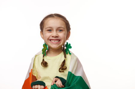 Close-up portrait of a cherry baby girl with clover leaves earring, smiling a toothy smile looking at camera, posing wrapped in flag of Ireland for Saint Patricks Day, on isolated white backgroundの写真素材