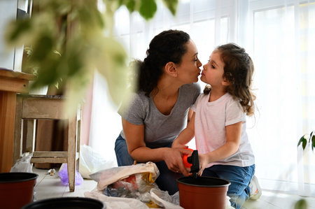 Loving mom kissing her daughter while taking care for houseplants. Little daughter learning gardening hobby in springtime, watering soil with plant seeds. Love and take care for planet from childhoodの写真素材