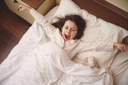 Top view of adorable little child girl in pajamas, lying down on a comfortable bed in children bedroom, outstretches her arms while waking up in the morning in happy mood. Kids. People. Childhoodの写真素材