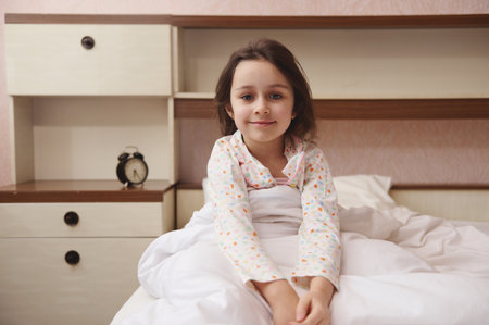 Lovely little child girl in pajamas, smiling looking at camera, sitting in bed at her cozy bedroom. Children. Lifestyle. Bedtime. Morning routines. The concept of going to bed, waking up and relaxingの写真素材