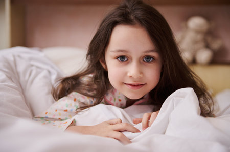 Close-up portrait of a lovely little child girl with long hair, looking at camera, lying in bed with...