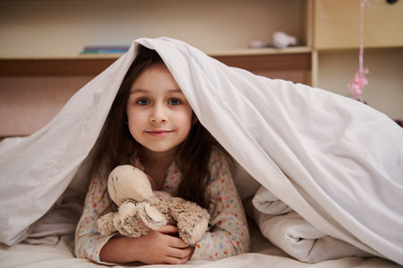 Portrait of lovely little child girl in pajamas, hugging her plush toy sheep, lying on her bed under comfortable soft white duvet, smiling looking at camera. Concept of childhood and daily routinesの写真素材