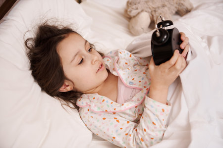 View from above of an adorable little girl in stylish pajamas with colorful dots, lying in bed with comfortable orthopedic mattress under cozy white duvet, starting alarm clock, getting ready for bedの写真素材