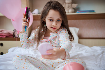 Beautiful kid girl sitting on her bed, looking at her reflection in a pink small mirror, taking care of her long hair, combing hair with colorful hair brush. Glamour kids. Beauty and body care conceptの写真素材
