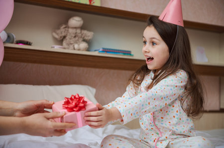 Amazed little child, birthday girl in pajamas, sitting on bed in her bedroom, expressing happy emotions while receiving a pink gift box with tied bow, with a happy surprise inside it for her birthdayの写真素材