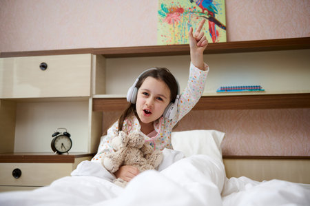 Adorable elementary aged child, baby girl in pajama and headphones, waking up in happy mood, holding a plush toy and enjoying soundtrack while sitting on her comfortable bed in light sunny bedroomの写真素材