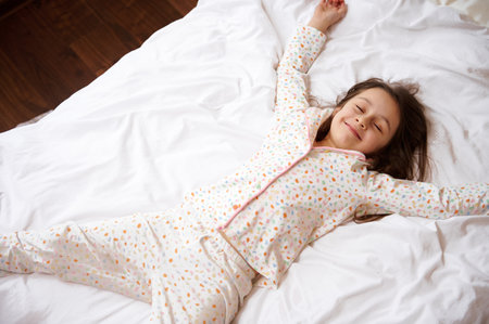 View from above of an adorable little child girl with closed eyes, in stylish cotton pajamas with colorful dots, smiling broadly while lying down on a comfortable bed with orthopedic mattressの写真素材