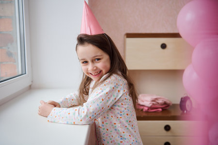 Little child, lovely birthday girl in pink party hat and white pajamas with colorful dots, standing by the window in her bedroom and smiles sweetly looking into camera, expressing positive emotionsの写真素材