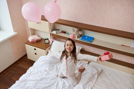 View from above: birthday little girl in party hat, holding bunch of pink pastel balloons and gift box, sitting on the bed in her cozy bedroom, expressing happiness, smiling cutely, looking at cameraの写真素材