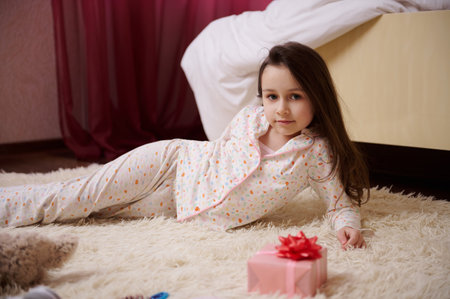 Adorable Caucasian little child girl with beautiful long hair, dressed in stylish pajamas with multicolored dots, lying on carpet in her bedroom, with a gift box on the foreground, looking at cameraの写真素材