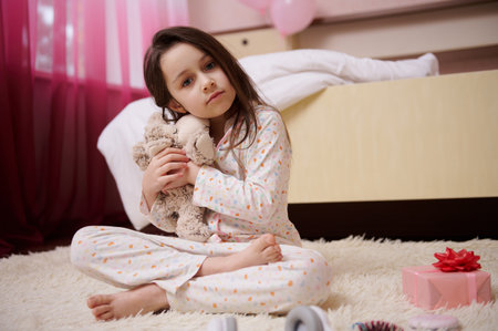 Adorable Caucasian child, lovely little girl in pajamas, hugging her plush toy sheep, looking at camera, sitting on the carpet in her room. International Childrens Day and happy childhood conceptの写真素材