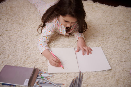 View from above of a Caucasian little child, a baby girl in stylish pajamas with multicolored dots, drawing picture with colorful pencils indoors. Art. Creativity. Children. Lifestyles. Leisure. Hobbyの写真素材