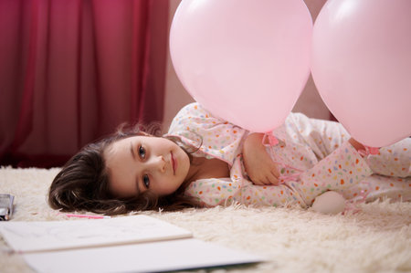 Caucasian adorable delightful little child girl in comfortable pajamas with multicolored dots, lying down on a carpet with thoughtful look in bedroom, playing with pink pastel balloons. Kids Childhoodの写真素材