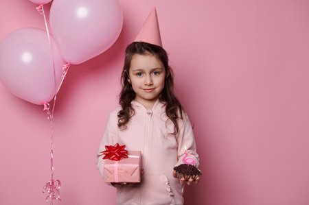 Beautiful happy little girl in pink clothes and party hat, holding a cute present for her birthday and a cupcake with candle, smiling looking at camera, isolated on pink background with air balloonsの写真素材