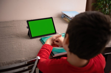 Rear view: smart student sitting a table a watching online lesson on a digital tablet with empty green chroma key screen with copy space for advertising text or mobile application. Distance educationの写真素材