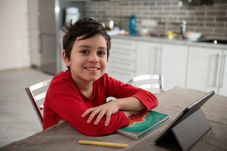 Confident smart preteen schoolboy, sitting with his arms folded at table with digital tablet, smiling broadly looking at camera. Adorable Caucasian teenage boy enjoys distance learning. Homeschoolingの写真素材