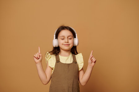 Lovely baby girl enjoys new sountrack, points her index fingers up and smiles, posing with her eyes closed on beige background with copy ad space. People. Children. Entertainment. Modern technologiesの写真素材