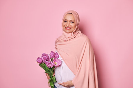 Isolated portrait on pink colored background of a Muslim Arab woman in pink hijab, holding a bouquet of purple tulips, touching her pregnant tummy, feeling happy by first baby kicks, smiling at cameraの写真素材
