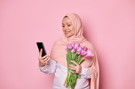 Middle-Eastern Muslim pregnant woman in pink hijab, using smartphone, scrolling news feed, checking social media content, testing new mobile app, posing with a bouquet of tulips, isolated backgroundの写真素材
