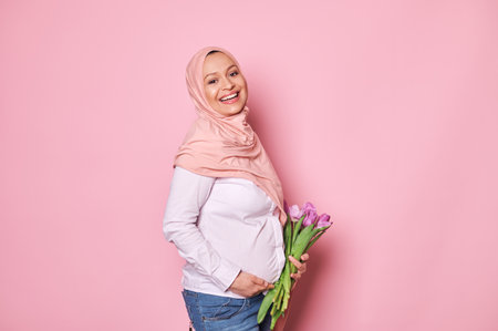 Delightful Muslim pregnant woman in pink hijab, smiling with beautiful toothy smile, looking at camera, touching her tummy, isolated pink background. Happy Mothers Day. Middle-Eastern female fashionの写真素材