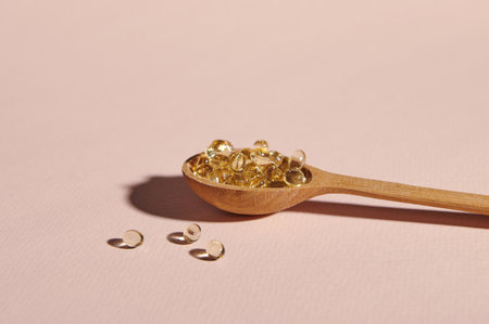Still life with wooden spoon full of soft gel capsules with golden color oil Omega 3 or vitamin D on pink pastel background with copy advertising space. Health care, medicine and pharmacy conceptの写真素材