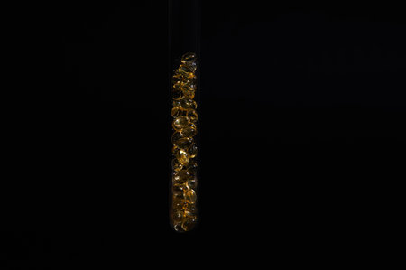 Still life with laboratory test tube filled with soft gel capsules with Omega 3 oil, vitamin D or essential acids, on isolated black background. Skin and body care. Beauty and pharmaceutical industryの写真素材