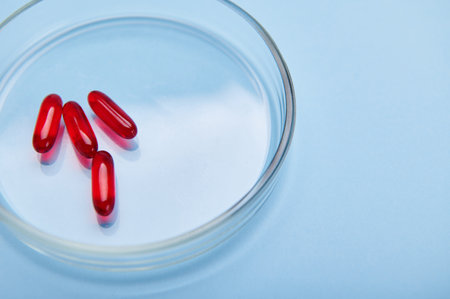 Overhead view of red gelatin translucent capsules with fat-soluble vitamin E, essential acid, Omega-3 or fish oil in Petri dish, isolated on blue color background with free ad space. World Health Dayの写真素材