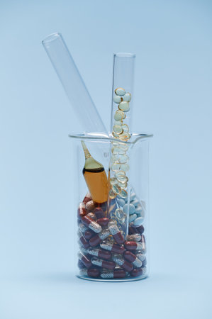 Vertical photo of a graduated laboratory beaker with pills, ampoule and test tubes with therapeutic dragees and translucent capsules, isolated on blue background. World Pharmacist and Health Day.の写真素材