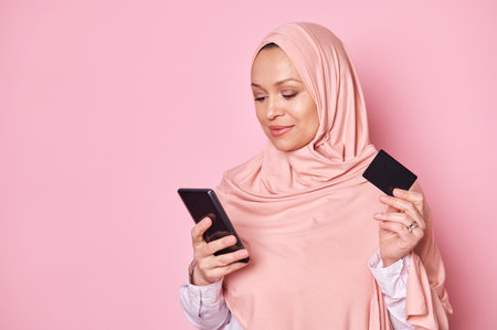 Closeup Beautiful young Arabic woman in pink hijab, using smartphone, shopping online, holding credit card, making payment via internet banking, isolated pink background. Mobile application E-Commerceの写真素材