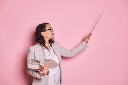 Confident pregnant woman, buisness coach, professor, teacher in stylish spectacles, conducting lesson, pointing to copy space with pointer, holding textbook in her hands, isolated over pink backgroundの写真素材