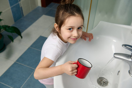 Mischievous baby girl standing by sink in home bathroom, holding red cup of water, smiling looking at camera while cleaning mouth, rinsing it with water while brushing teeth. Dental care. Oral hygieneの写真素材