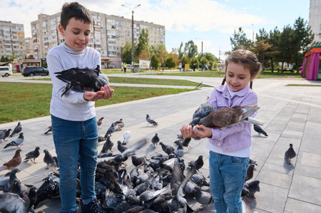 Two adorable kids, brother and sister feed pigeons in the park square. Spring. Lifestyle. The concept of kindness, care for animals. Family outing. A walk with benefit. Children, nature and animals.の写真素材