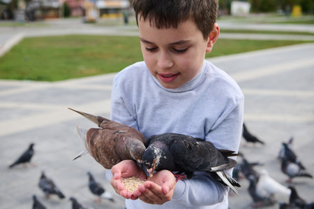 Close-up portrait of Caucasian adorable teen boy having fun during family outing, feeding birds from his hands in park square. The concept of kindness, walking with benefits, love and care for animalsの写真素材