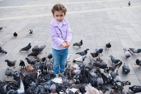Full-length portrait of a Caucasian lovely little girl dressed in casual denim, enjoying a family outing with benefits, feeding rock doves crowding the street, and eating discarded food and bird seedsの写真素材
