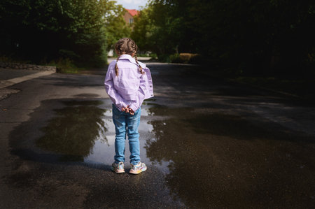 Little girl in jeans and purple jacket, standing with her hands crossed by a small puddle after rain and looking at her reflection in the water while walking on the countryside street. Rear viewの写真素材