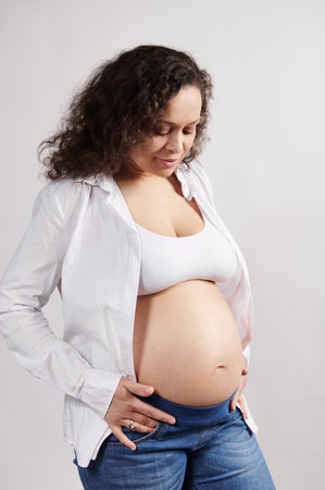 Vertical studio portrait. Beautiful curly haired multi ethnic pregnant woman in blue jeans, looking with tenderness and love at her naked belly, isolated white background. Pregnancy 6 month. 24 weeksの写真素材